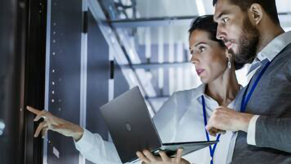A man holding a laptop standing next to a woman pointing at a control panel