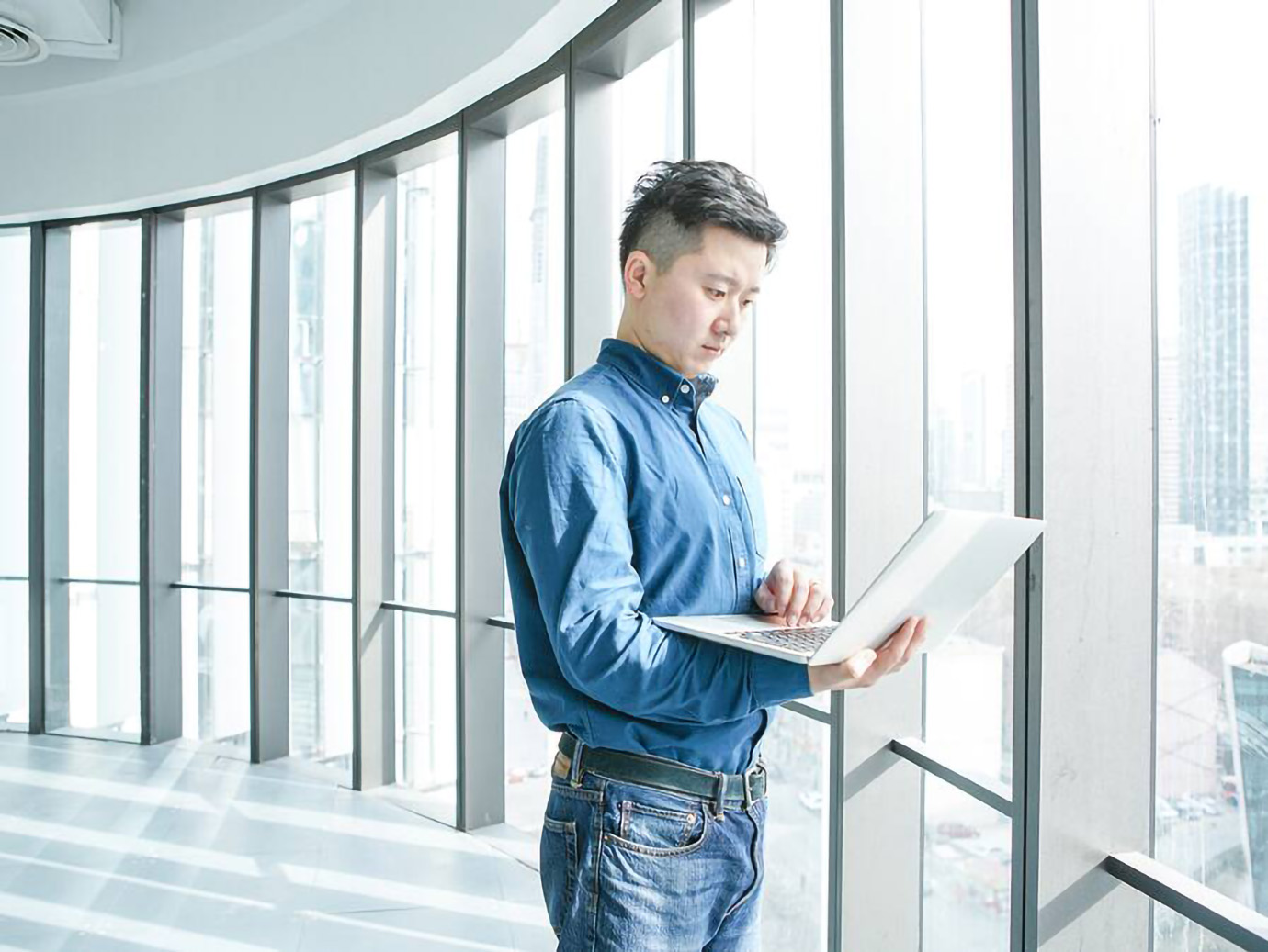 Man holding a laptop in his hand and standing next to a glass window in a commercial building