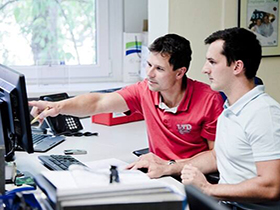 A man is pointing to a computer monitor and showing something to another man
