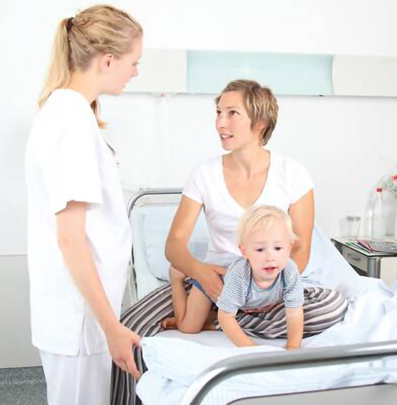 A nurse speaking to a mother and child seated on the hospital bed
