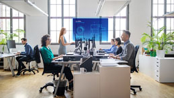 Employees sitting at a table watching a presentation with the OpenBlue Enterprise Manager ​overlaid on the screen