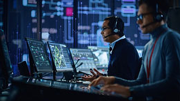 Two men wearing headsets working on computers in a control room